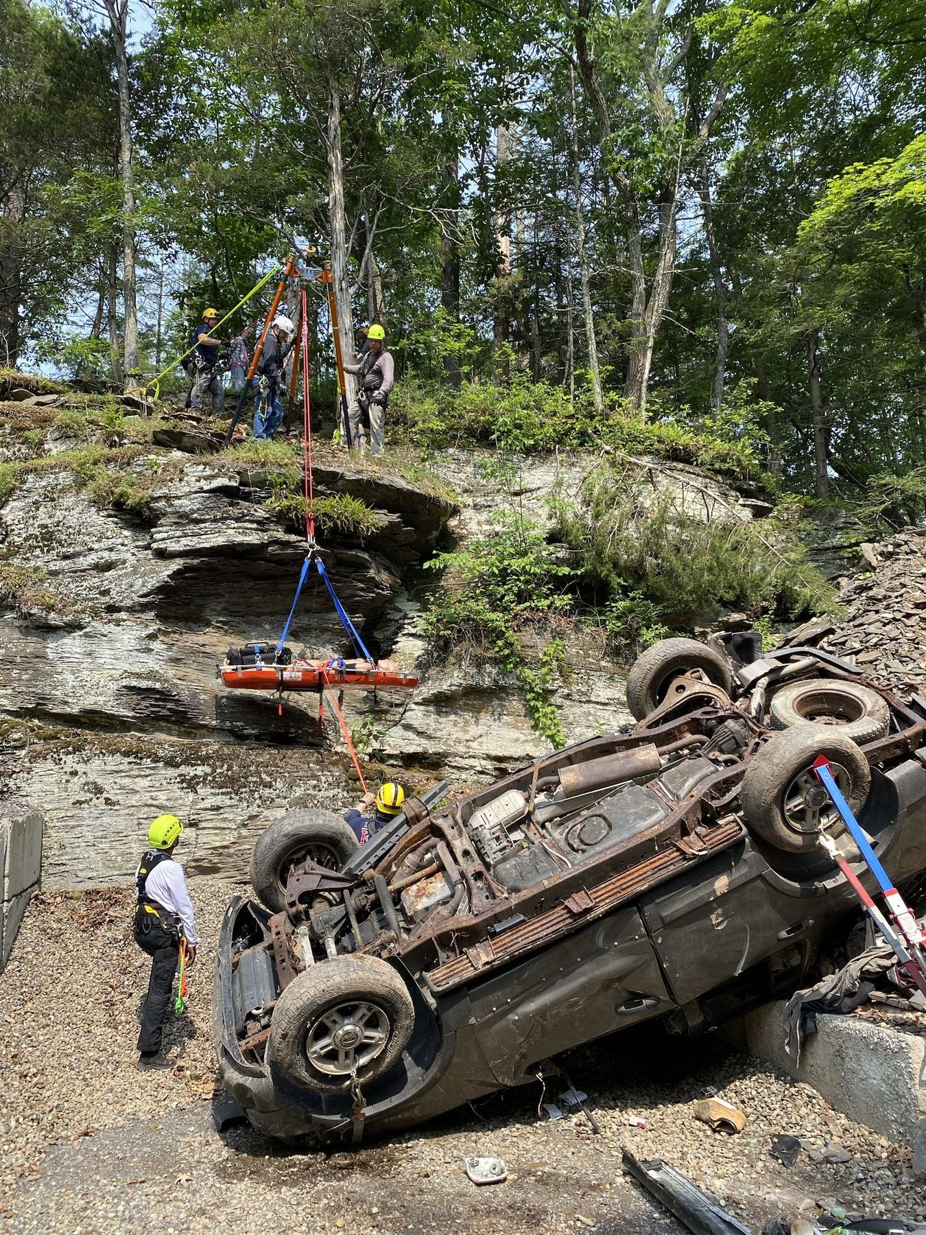 A patient in a basket litter being lowered down a rock face past an overturned pickup