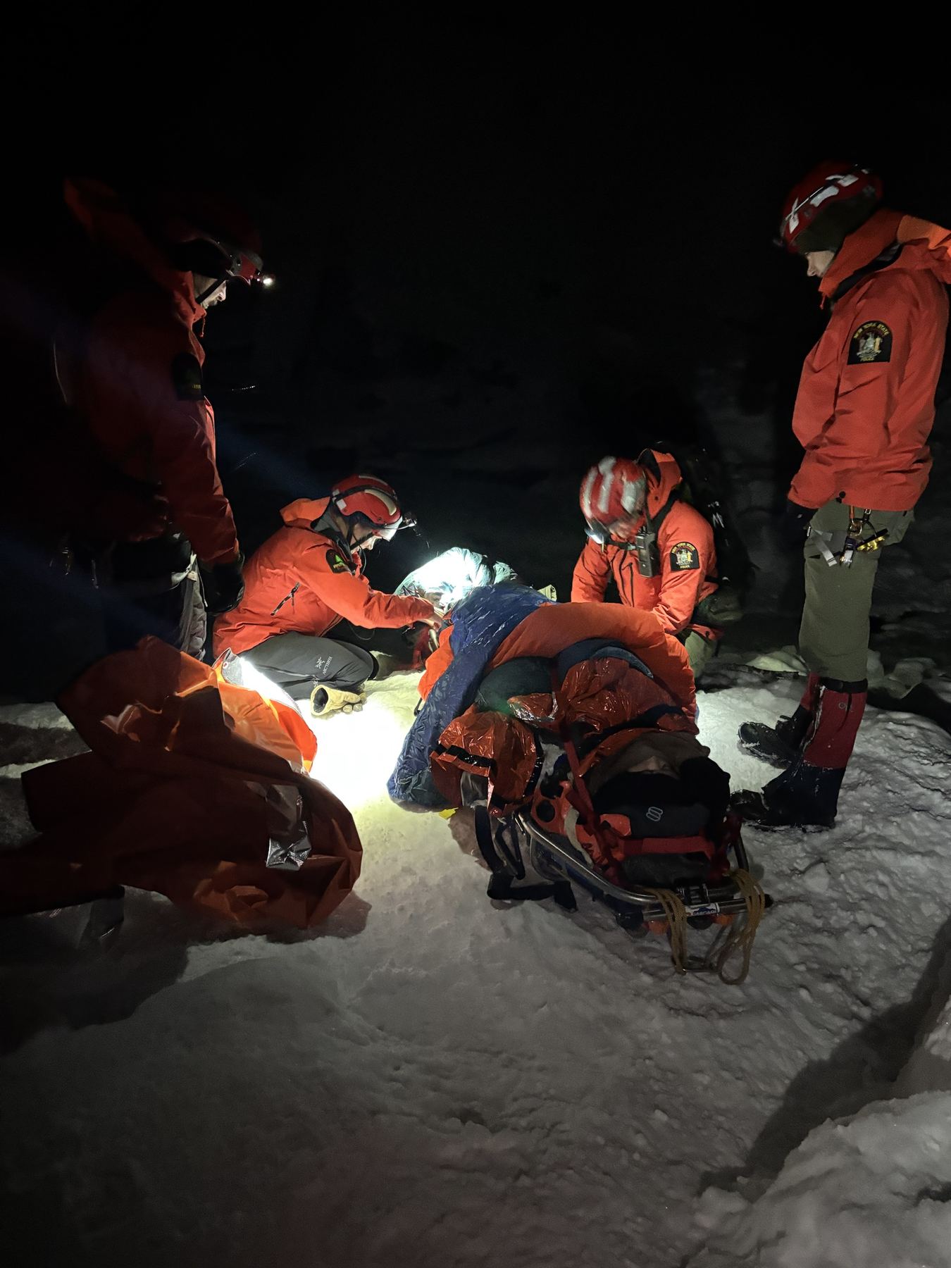 Rescuers in red jackets and helmets packaging a patient in a litter on snow at night
